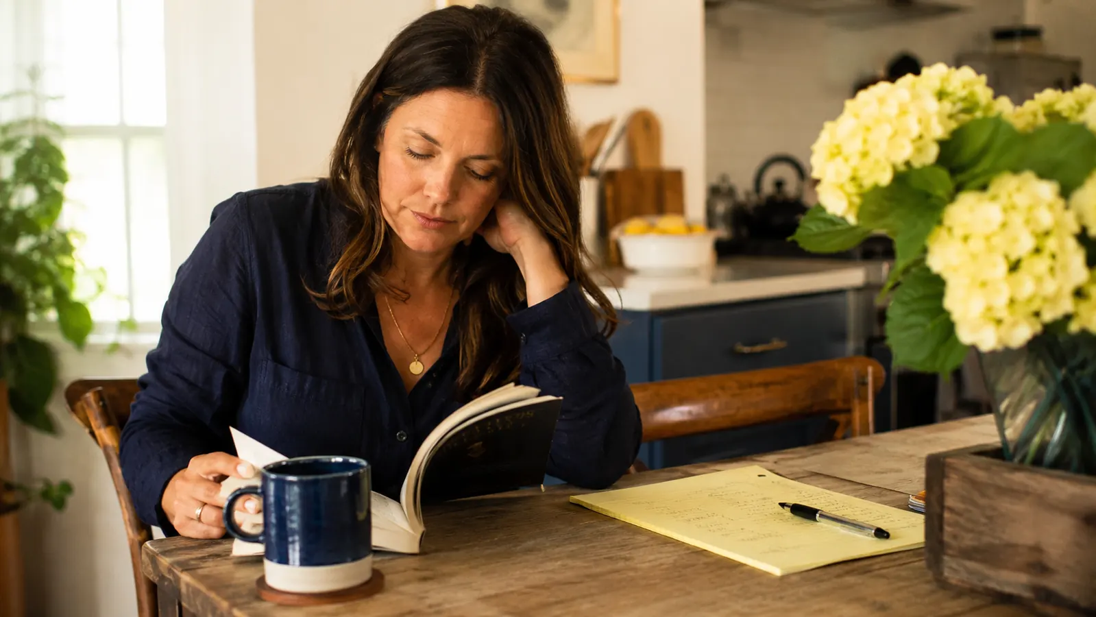 Woman sitting at a kitchen table with a personal finance book open next to a cup of coffee and a notepad with budget figures